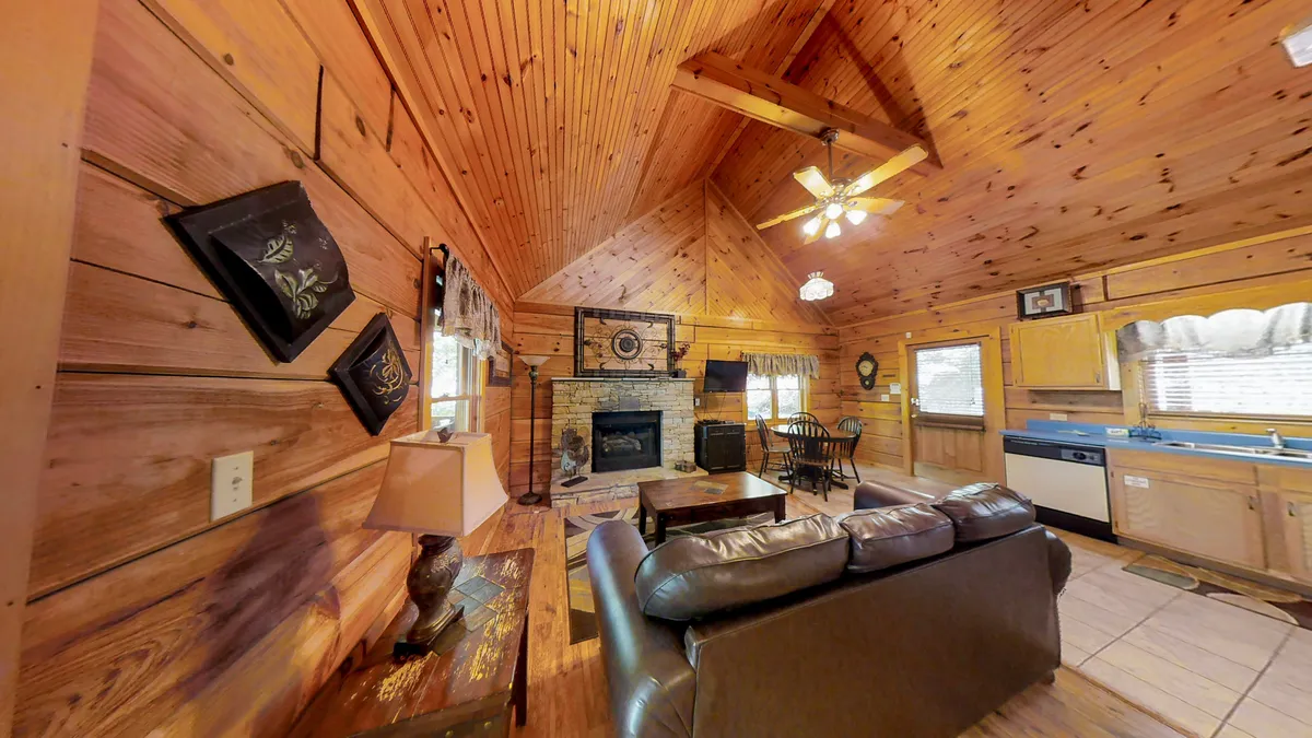 A warmly lit living area featuring a comfortable sofa set near a seasonal gas fireplace, wooden beams overhead, and a visible dining area in the background. The space exudes a cozy, homey feel, with inviting textures and a seamless transition into the dining zone.