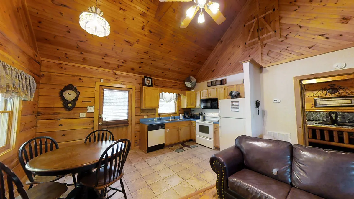 A bright and welcoming open-plan space showing the dining table, a comfortable sofa, a glimpse of the kitchen area, and a wooden ceiling overhead. The airy atmosphere and natural light highlight the seamless flow between living, dining, and kitchen spaces.