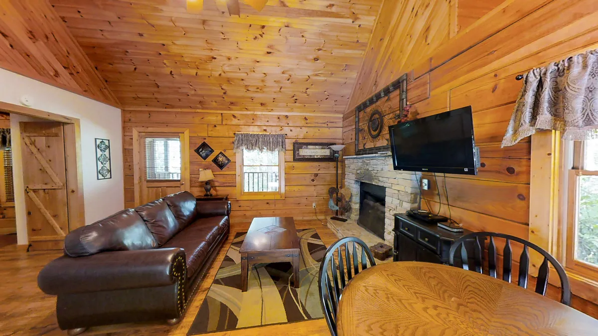 A spacious, inviting living room with a brown leather sofa facing a stone fireplace, a cozy wooden coffee table, and an adjacent dining table. The scene is perfect for relaxing, enjoying meals, and spending time with family or friends.