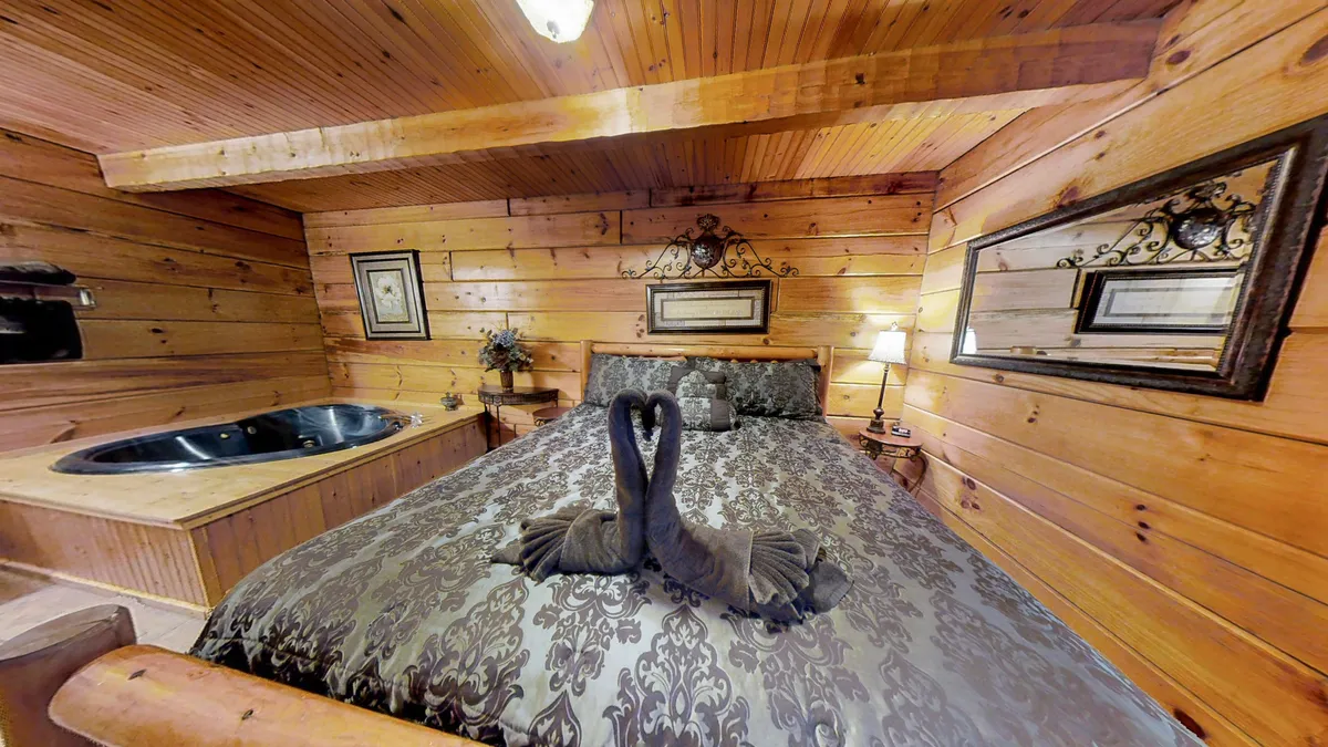 A warmly lit master suite with a king bed adorned in decorative bedding, swans crafted from towels atop the bed, and a heart-shaped Jacuzzi tub in the corner. The wooden paneling and intimate layout create a romantic and inviting ambiance ideal for couples.